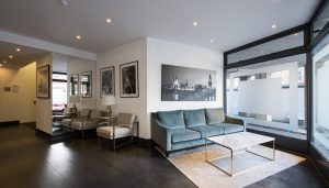 A modern, minimalist lobby features a teal sofa, marble coffee table, and artwork depicting cityscapes. Mirrored walls reflect seating and enhance the sense of space.