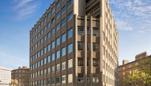 Luke House, a large, multi-story building with repeating square windows, is showcased. The stone building stands tall against a bright blue sky.
