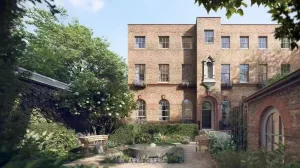 A brick building with multiple windows and a central entrance, surrounded by trees and shrubs. A small pond is visible in the foreground, along with seating areas and pathways in a garden setting.