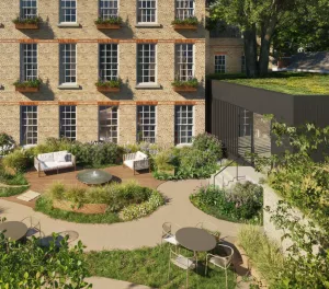 A landscaped garden area featuring seating arrangements, a round table, and various plants. The background shows a brick building with windows and flower boxes, alongside a modern structure with a green roof.