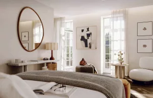 A cozy bedroom featuring a bed with gray bedding, a round mirror on the wall, and large windows letting in natural light. A small side table with a lamp and a modern chair are also visible.