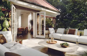 A modern outdoor patio with a white sectional sofa and a round coffee table. Glass doors open into a well-lit interior with dining furniture. Lush green plants are visible, and sunlight filters through the trees.