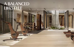 An indoor fitness area featuring exercise bikes and strength training equipment. Large windows show greenery outside, and a tree is visible in the center of the gym. The floor is wooden and the decor is modern.